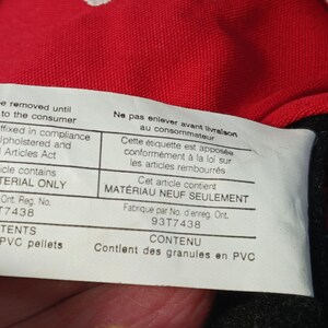 Op de afbeelding: Close-up van een wit label met zwarte tekst, bevestigd aan rode stof. Het label vermeldt "Niet verwijderen tot levering aan de consument" en "Dit artikel bevat ALLEEN NIEUW MATERIAAL". Het label bevat ook de tekst "Bevat PVC-korrels".