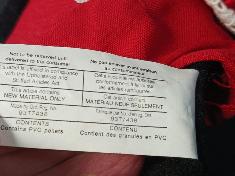 Op de afbeelding: Close-up van een wit label met zwarte tekst, bevestigd aan rode stof. Het label vermeldt "Niet verwijderen tot levering aan de consument" en "Dit artikel bevat ALLEEN NIEUW MATERIAAL". Het label bevat ook de tekst "Bevat PVC-korrels".