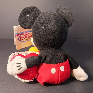 Op de afbeelding: Een Mickey Mouse pluche speelgoed, gezien van achteren, met een rood hartvormig kussen met witte kanten rand. Mickey draagt een rode broek met witte knopen en witte handschoenen. Het Disney Store-label is zichtbaar.