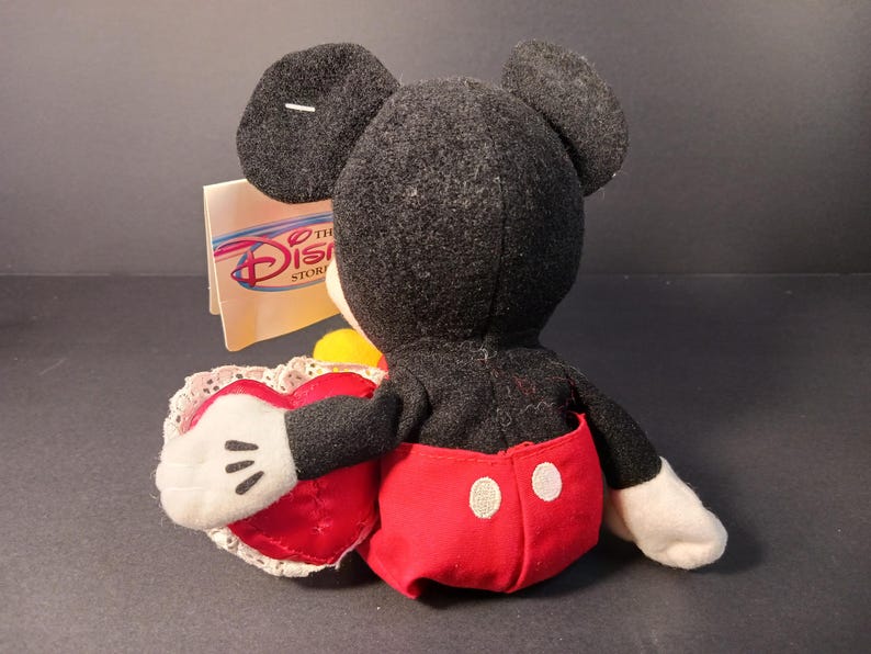 Op de afbeelding: Een Mickey Mouse pluche speelgoed, gezien van achteren, met een rood hartvormig kussen met witte kanten rand. Mickey draagt een rode broek met witte knopen en witte handschoenen. Het Disney Store-label is zichtbaar.