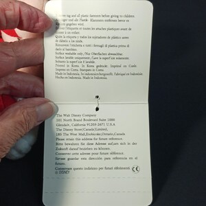 Op de afbeelding: Een wit productlabel met tekst in meerdere talen, waaronder Engels, Frans en Spaans. Het label wordt door een hand vastgehouden en de tekst bevat onderhoudsinstructies en bedrijfsinformatie.