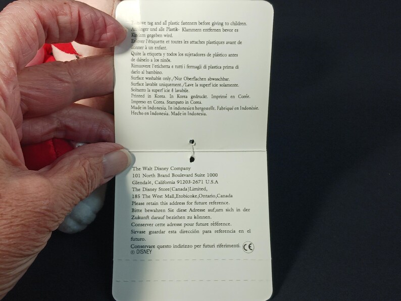 Op de afbeelding: Een wit productlabel met tekst in meerdere talen, waaronder Engels, Frans en Spaans. Het label wordt door een hand vastgehouden en de tekst bevat onderhoudsinstructies en bedrijfsinformatie.