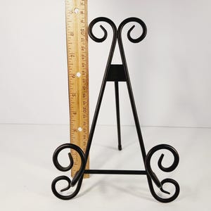 Op de afbeelding: Zwarte metalen display-ezel met decoratief krulwerk aan de boven- en onderkant. De ezel is ongeveer 30 cm hoog en heeft een driepootontwerp. Een houten liniaal wordt naast de ezel geplaatst.