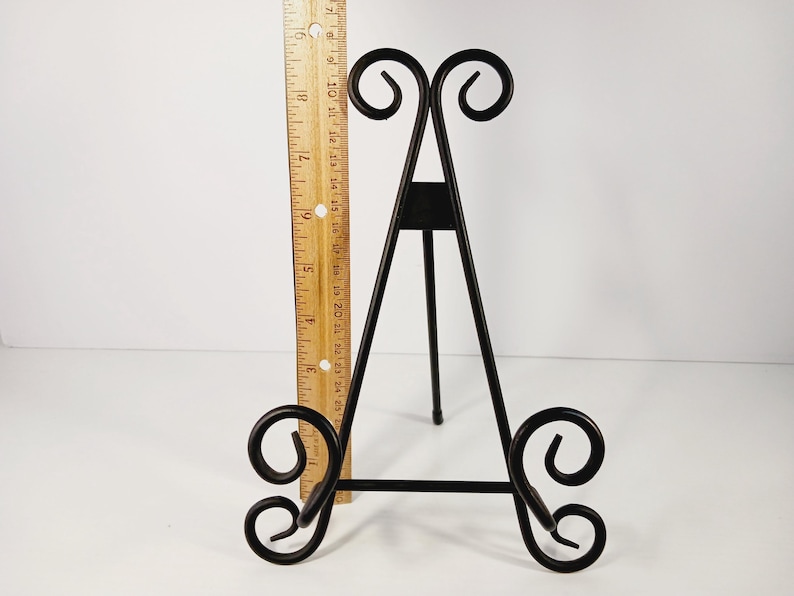 Op de afbeelding: Zwarte metalen display-ezel met decoratief krulwerk aan de boven- en onderkant. De ezel is ongeveer 30 cm hoog en heeft een driepootontwerp. Een houten liniaal wordt naast de ezel geplaatst.