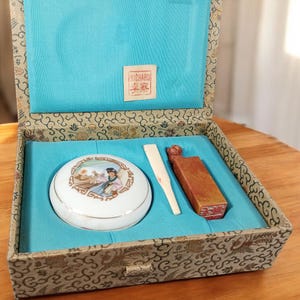 May include: An open decorative box with a blue interior, containing a white porcelain container with a painted scene, a small wooden fan, and a rectangular, reddish-brown object. The box has a patterned exterior and a small label.