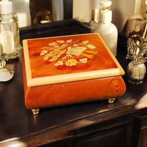 May include: A wooden music box with a decorative inlaid design on the lid. The lid features a violin, sheet music, and floral accents. The box is rectangular with rounded edges and rests on small, gold-colored feet.