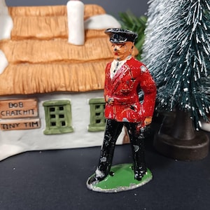 May include: A painted metal figurine of a standing figure wearing a red jacket, black trousers, and a black cap. The figure stands on a green oval base. In the background, a ceramic building features signs reading Bob Cratchit and Tiny Tim.