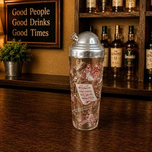 May include: A vintage cocktail shaker with a silver lid and a clear glass body. The glass is decorated with red illustrations and text. The shaker is on a dark wooden surface, with bottles of liquor in the background. A sign reads "Good People Good Drinks Good Times."