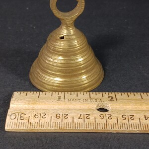 Puede incluir: Una peque&ntilde;a campana de lat&oacute;n con un asa en forma de lazo. La campana tiene forma c&oacute;nica con crestas horizontales. Una regla de madera, marcada en pulgadas, se coloca junto a la campana. La campana mide unos 7,6 cm de alto.