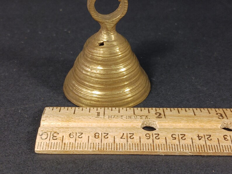 Puede incluir: Una peque&ntilde;a campana de lat&oacute;n con un asa en forma de lazo. La campana tiene forma c&oacute;nica con crestas horizontales. Una regla de madera, marcada en pulgadas, se coloca junto a la campana. La campana mide unos 7,6 cm de alto.