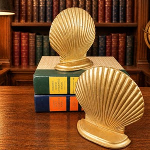 May include: Two gold-colored seashell bookends, one upright and one on its side, rest on a wooden surface. They are positioned in front of a stack of books with green and blue covers. A library setting with a globe and books in the background.