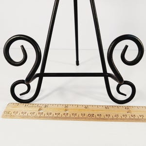 Op de afbeelding: Zwart metalen ezel met decoratief krulwerk op de basis. De ezel heeft drie poten en een verticale steun. Een houten liniaal is voor de ezel geplaatst. De liniaal meet tot 30 cm.