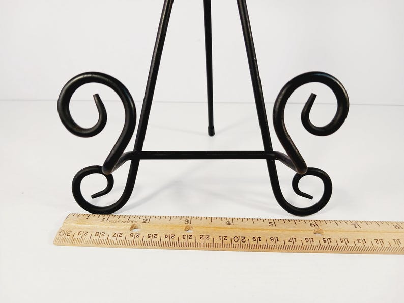 Op de afbeelding: Zwart metalen ezel met decoratief krulwerk op de basis. De ezel heeft drie poten en een verticale steun. Een houten liniaal is voor de ezel geplaatst. De liniaal meet tot 30 cm.