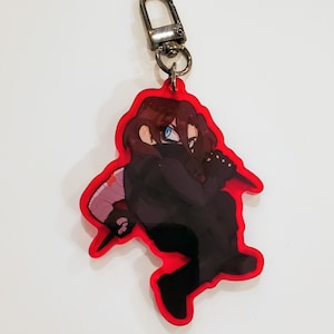 Puede incluir: Un llavero con contorno rojo que presenta un personaje de estilo anime con un atuendo negro, cabello castaño y ojos azules. El llavero tiene un cierre plateado.