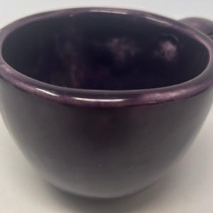 May include: A small, deep purple ceramic cup with a smooth, glossy finish and a rounded, bowl-like shape. The cup features a thick, curved rim and a single handle extending from the side, showcasing a uniform dark violet colour throughout the exterior.
