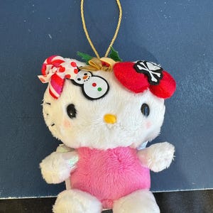 May include: A plush white Hello Kitty ornament with a pink dress and a red bow with a skull and crossbones. The ornament has a gold string for hanging and a snowman design on the other side of the head.