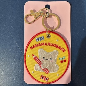 May include: A yellow embroidered keychain with a red border and gold-colored hardware. The keychain features a cartoon bear holding a red pencil, with the text "HANAMARUOBAKE" in a circular design.