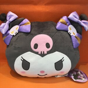 May include: A plush, gray character pillow with a pink skull face and black eyes. The pillow has purple bows with skull accents and gold trim. The character's face is white with a pink nose and a mischievous expression. A tag is attached to the pillow.