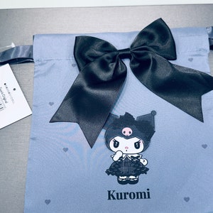 May include: A light blue drawstring bag with a black satin bow and a black and white cartoon character with the name "Kuromi" printed on the front. The bag has a white tag with black text and a barcode.