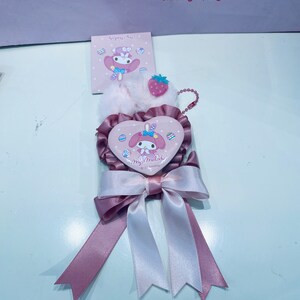 May include: Pink and white heart-shaped charm with a My Melody character design. The charm is attached to a pink and white ribbon bow. The charm has a small strawberry charm attached to it.
