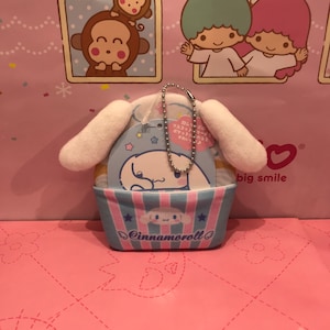 May include: A white Cinnamoroll plush keychain with a blue and pink striped basket. The keychain has a silver chain and a tag that says "Cinnamoroll" and has Japanese text.