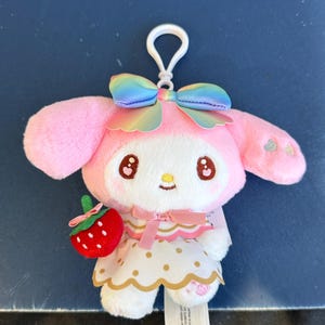 May include: A pink plush toy with a white face, brown eyes, and a rainbow bow. The toy has long ears, a white and gold dress, and a red strawberry accessory. A silver keychain is attached to the top.