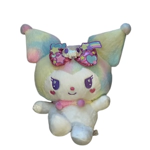 May include: A plush toy featuring a white body with a pastel rainbow-colored head and ears. The toy has a decorative bow with stars and a skull. It has purple eyes, pink cheeks, and a pink scarf.