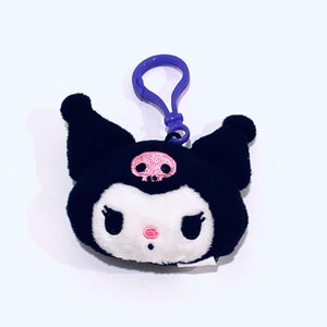 May include: A plush black character with a white face, pink nose, and a pink skull on its forehead. It has a purple clip attached. The character has large black eyes and black ears. The plush is on a white background.