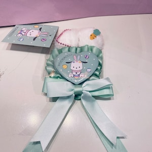 May include: A green heart-shaped pin with a white ribbon. The pin features a cartoon dog with a pink bow and a carrot. The text "POCHACCO" is printed on the pin.