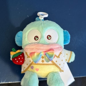 May include: A plush toy in the shape of a cartoon character, primarily in shades of teal, pink, and yellow. It features large eyes, a rainbow bow, and a red strawberry. The toy has a small loop at the top for hanging.