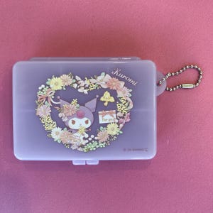 May include: A purple plastic pill box with a floral heart design featuring a cartoon character named Kuromi. The box has a small metal chain attached to it. The text "Kuromi" and "For you" are printed on the box.
