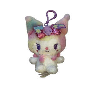 May include: A plush toy featuring a white body, pink and rainbow ears, and purple eyes. It has a pink bow with a skull and a purple keychain. The toy is a cute, kawaii-style character.