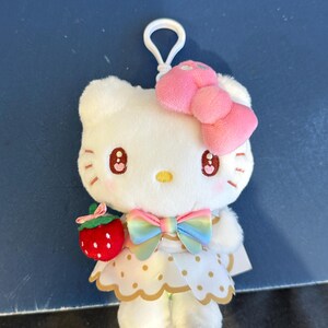 May include: A white plush toy featuring a cat-like character with a pink bow, a rainbow bow, and a strawberry accessory. The character has a white dress with gold polka dots and a white clip.
