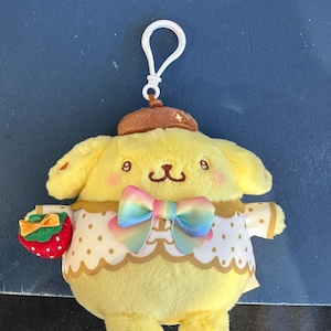 May include: A yellow plush keychain featuring a cartoon dog character with a brown hat and a rainbow bow tie. The dog is holding a small strawberry with a green bow. The keychain has a white loop.