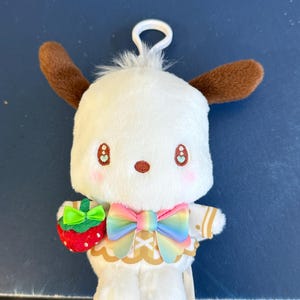 May include: A white plush toy with brown ears, holding a red strawberry with a green bow. The toy has a rainbow bow and a gold-trimmed outfit. It has a white loop for hanging.