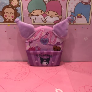 May include: Purple plush cupcake-shaped coin purse with a pink and white swirl design, a cherry, and a chocolate chip cookie. The purse has a pink heart with glitter and the text "Kuromi" in pink letters.