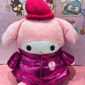 May include: Pink plush toy with a pink and white hat, wearing a pink metallic jacket with a white bow on the front. The toy has a white face with black eyes and a yellow nose.