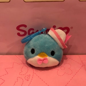 May include: A plush blue bird-shaped keychain with a white and pink sailor hat and a pink bow tie. The bird has black eyes and a yellow beak. A blue plastic clip is attached to the top.