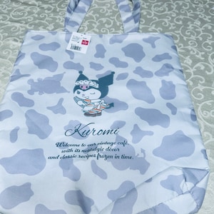 May include: A white tote bag with a cow print pattern. The bag features a cartoon character named Kuromi, who is wearing a black dress and a crown. The text "Kuromi" is written in a blue font above the character. The text "Welcome to our vintage café, with its nostalgic décor and classic recipes frozen in time." is written in a blue font below the character.