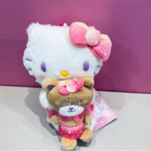 May include: A white plush Hello Kitty with a pink bow and a smaller brown teddy bear wearing a pink outfit. The teddy bear is held by Hello Kitty. Both toys have pink accents and are set against a purple background.