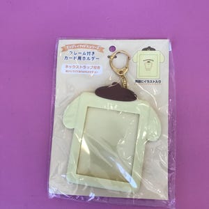 May include: A light green plastic card holder shaped like a cartoon dog wearing a brown shirt. The holder has a clear plastic window for inserting a card and a gold metal key ring attached.
