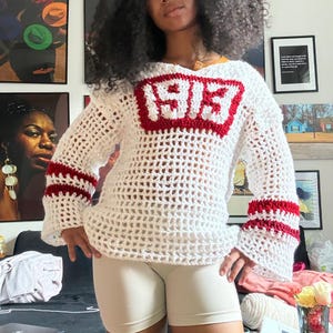 May include: White crochet sweater with red accents. The sweater features a red block with the numbers "1915" on the front and red stripes on the sleeves. The sweater has a loose, open weave design.