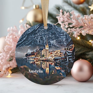 May include: A round, glass bauble featuring a painted image of a small Austrian village. The village is situated in a snowy mountain valley with a lake in the foreground. The bauble is decorated with the word "Austria" at the bottom.