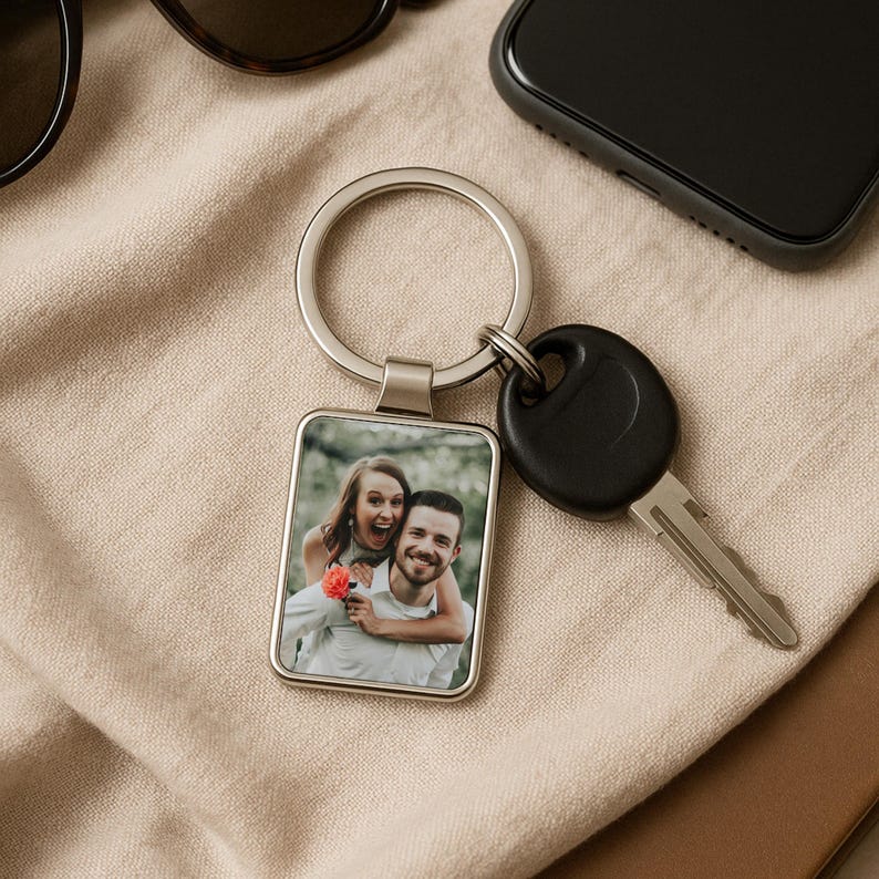 May include: A silver keyring with a rectangular photo charm featuring a couple smiling. The photo charm is attached to a silver key ring and a black key.