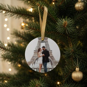 Puede incluir: Un adorno navide&ntilde;o redondo de cer&aacute;mica blanca con una fotograf&iacute;a impresa de una pareja frente a la Torre Eiffel. El adorno est&aacute; suspendido por una cinta dorada y cuelga de una rama de pino verde con suaves luces bokeh de fondo.