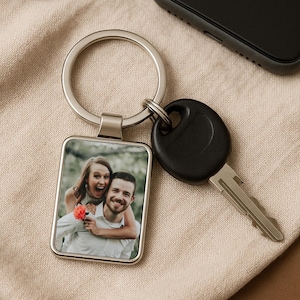 May include: Silver rectangular keychain with a photo of a smiling couple. The keychain is attached to a key and rests on a beige fabric surface. A black smartphone is in the background.