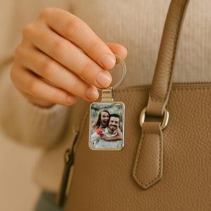 May include: A silver keyring with a rectangular photo insert featuring a couple smiling. The photo shows a woman with long brown hair wearing a white shirt and a man with short brown hair wearing a blue shirt. The keyring is attached to a brown leather handbag.