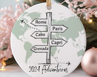 Puede incluir: Adorno redondo blanco con un mapa del mundo verde y un poste indicador con flechas que apuntan a Roma, París, Cabo, Capri y Granada. El poste indicador tiene el texto "2024 Adventures" debajo.