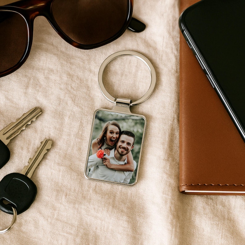 May include: A silver keyring with a rectangular photo insert featuring a couple smiling. The photo shows a woman with long brown hair wearing a white shirt and a man with short brown hair wearing a white shirt. The woman is holding a pink flower.