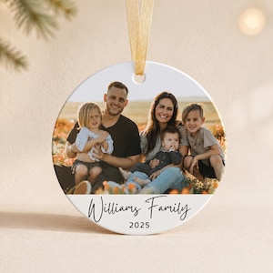 Puede incluir: Un adorno redondo de cer&aacute;mica blanca con una fotograf&iacute;a de una familia de cuatro personas sentadas en un campo. El texto "Williams Family" est&aacute; impreso en negro bajo la foto, con el a&ntilde;o "2025" debajo. Una cinta dorada est&aacute; sujeta en la parte superior.